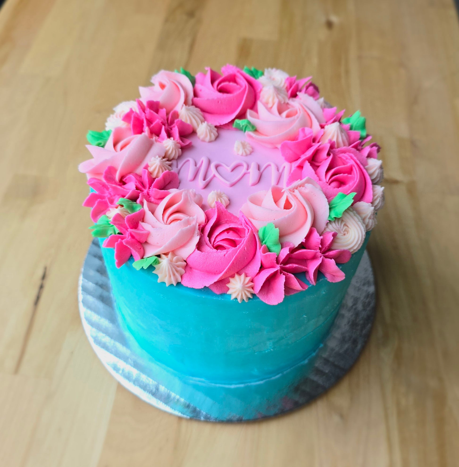 A bright floral-themed cake available during a Mother's Day workshop, with blue frosting on the sides, and on the top: pink rosettes, green leaves, and mom written in the center. 