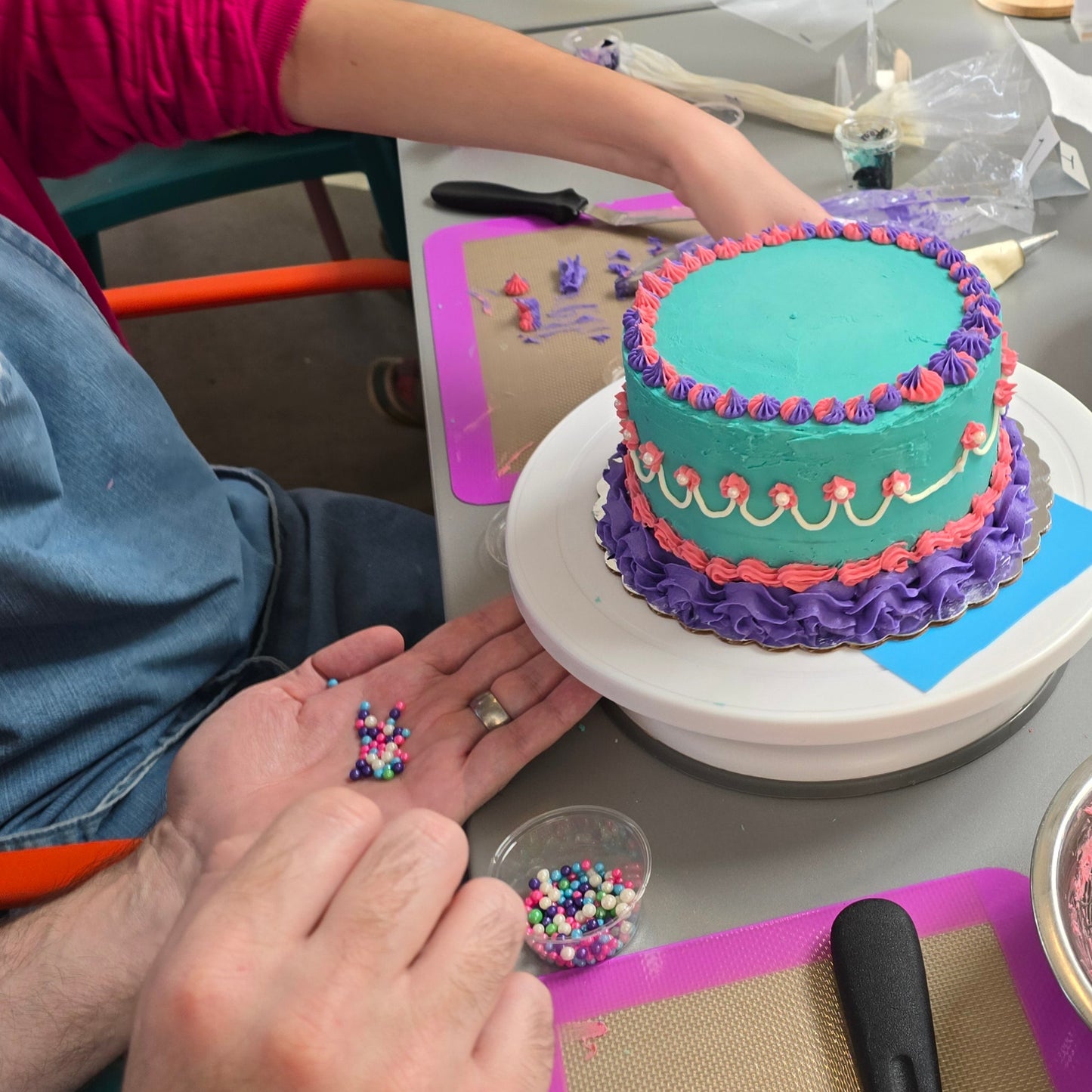 A couple works on a a purple, teal, and pink cake decorated with piped borders, shells, and writing at Cake Hoopla in Tigard, Oregon, near Portland, during a cake-decorating class.