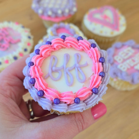 Cupcake with 'bff' frosting held by a hand in front of other similar cupcakes in the background.