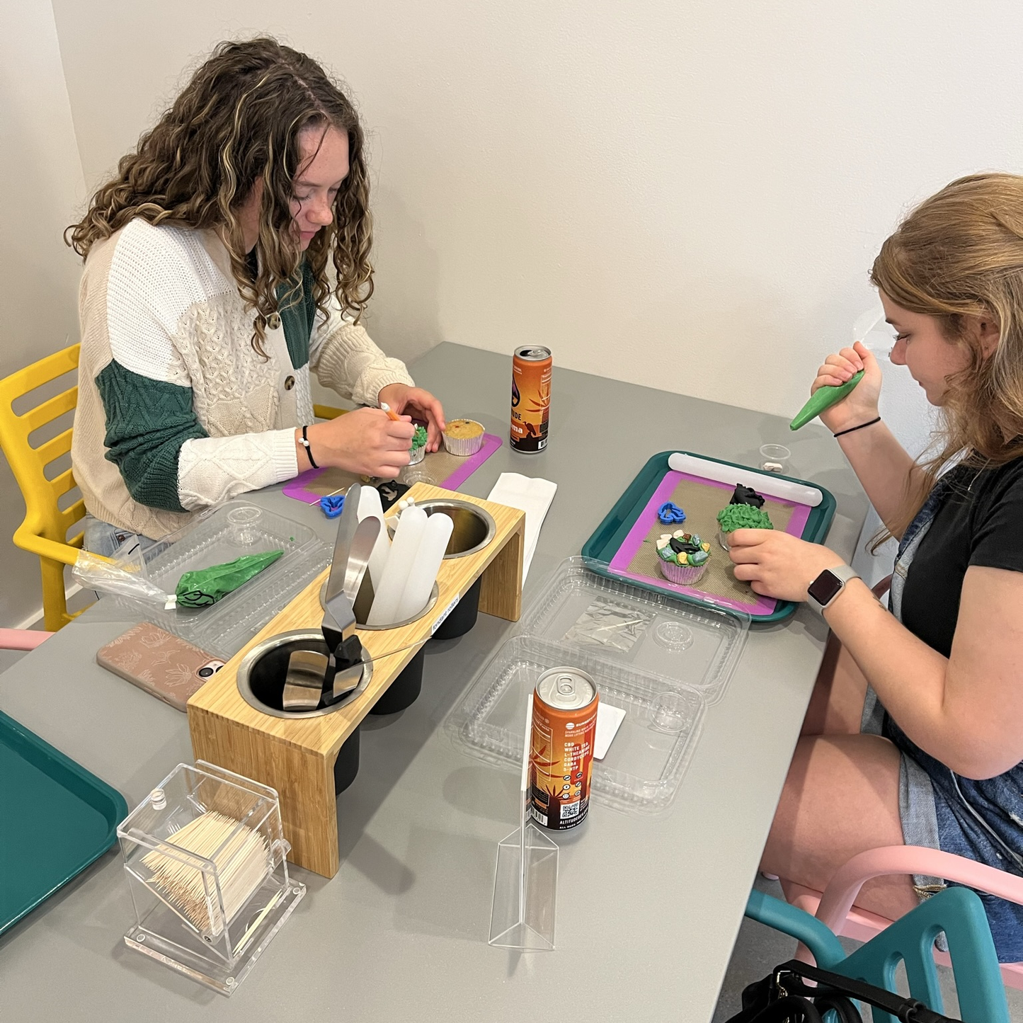 Two young ladies decorate cupcakes with frosting and tools, while sipping a canned beverage, in the Cake Hoopla party room for a DIY cupcake-decorating event in Tigard, near Portland.