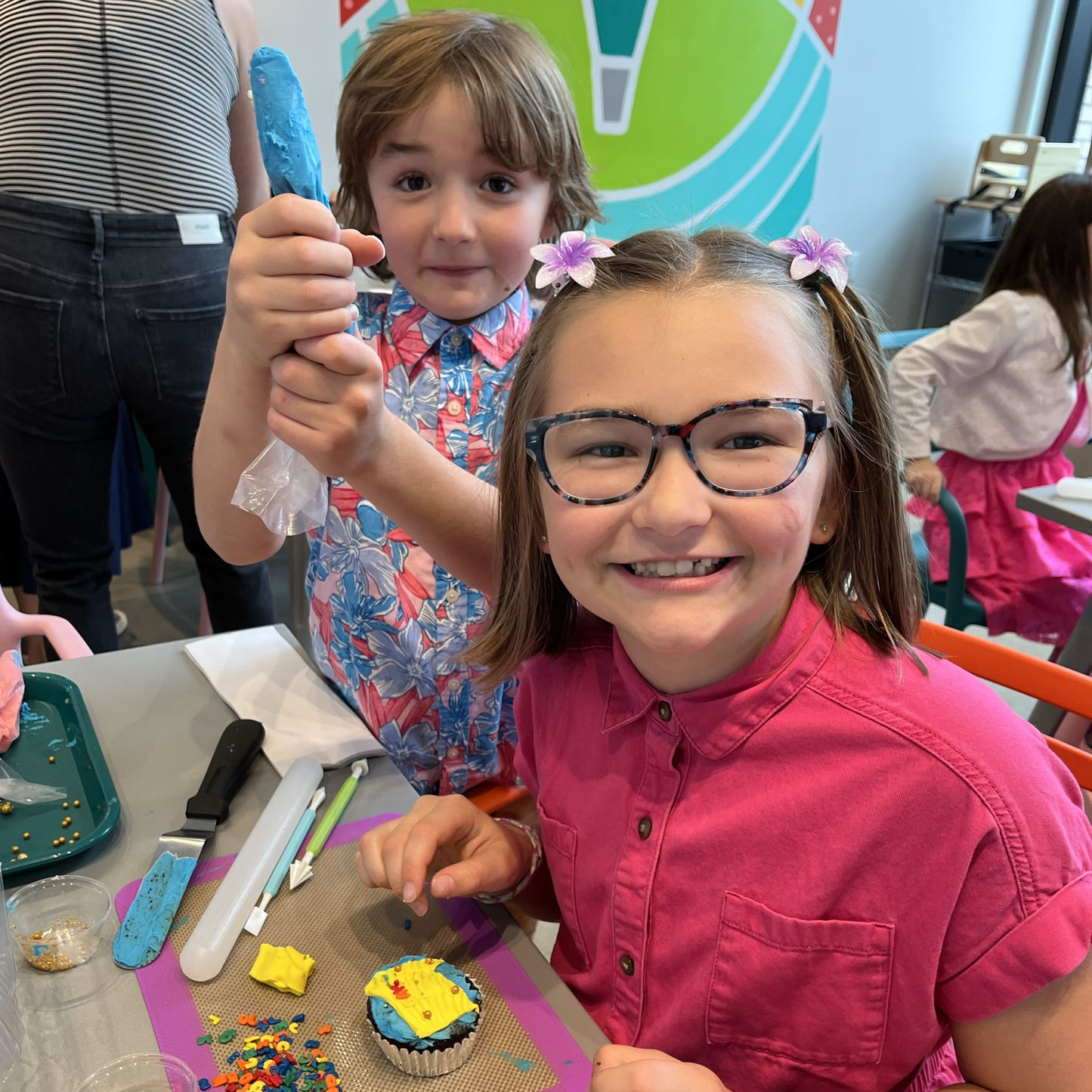 At Cake Hoopla's DIY cake/cupcake-decorating studio, two elementary-aged kids show off their cupcake-decorating skills with a piping bag of blue frosting and a frosted cupcake with a yellow fondant and sprinkle design.
