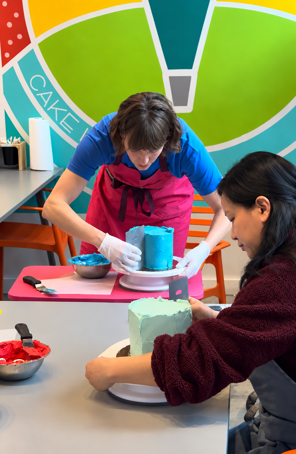 A cake instructor teaches how to frost a blue heart-shaped cake using a metal scraper with the Cake Hoopla studio in the background.