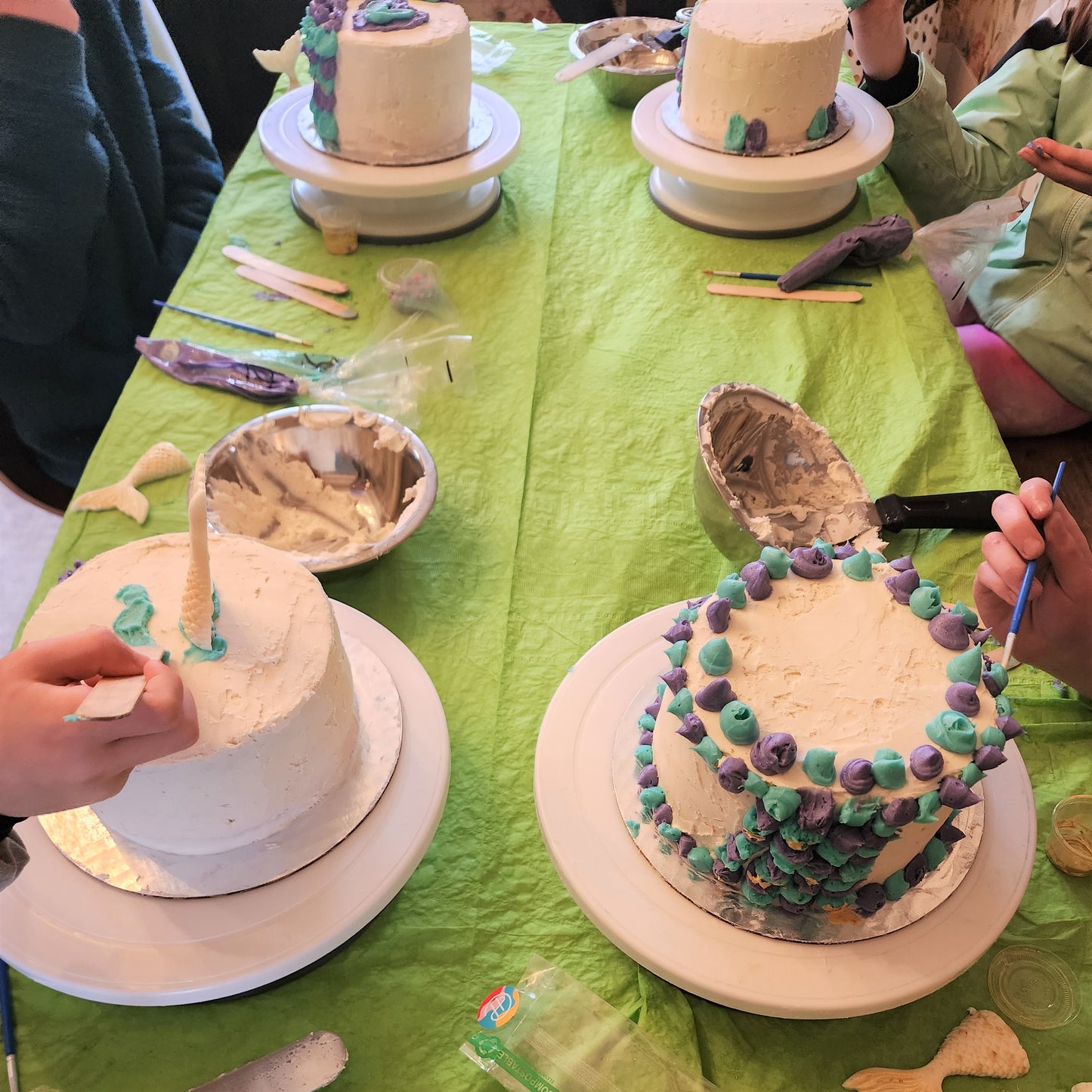 kids are decorating four cakes in the theme of a mermaid with teal and purple frosting scales and a white chocolate mermaid tail on top of the cakes for a DIY cake-decorating party at Cake Hoopla in Tigard, near Portland.