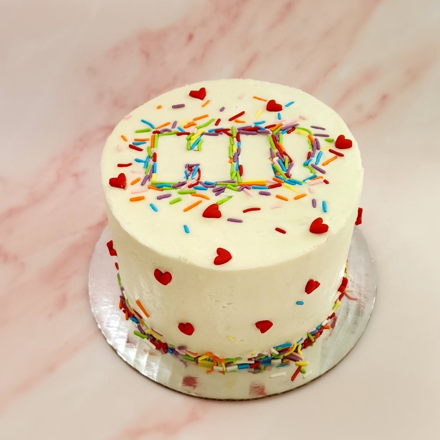 A decorated DIY Just Hi cake. White frosted cake decorated with rainbow sprinkles forming the word "Hi" in the center and cute red fondant hearts dotted around the front and sides of the cake.
