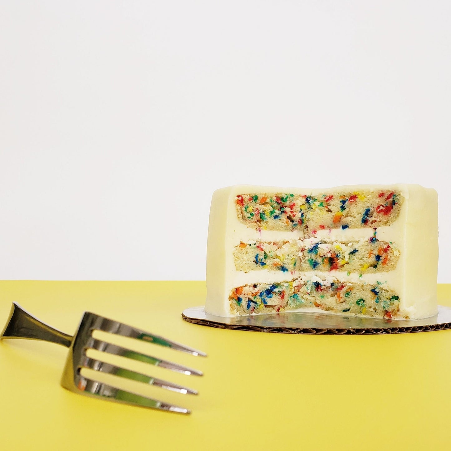 Cake Hoopla’s “Fetti” cake, cut open to reveal the inside layers. A three-layer vanilla cake has tons of bright-colored confetti sprinkles baked into the layers, and white vanilla buttercream frosting separating each layer. The cake looks moist and delicious.