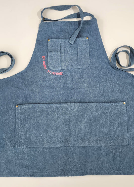 A medium-blue denim apron with a utility pocket across the breast. Tracing the bottom corner of the pocket are the words "go cake yourself" in neon pink embroidery. Across the lower body of the apron is a large open pocket. Small bras rivets stamp the upper corners of each pocket. A denim neck strap is secured with a brass-colored loop buckle. 2 long straps rest on either side of the apron. This apron is modern with denim a slight Americana feel.