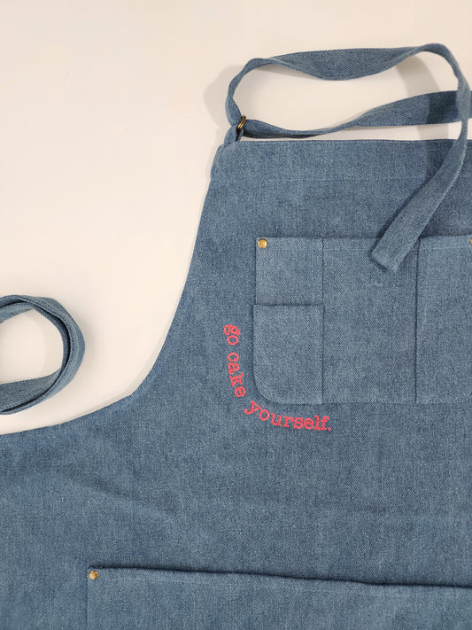 A closeup view of the corner of the breast pocket outlined with neon pink embroidery reading "go cake yourself." The font has a typewritten look and is somewhat discrete for the size of the apron.