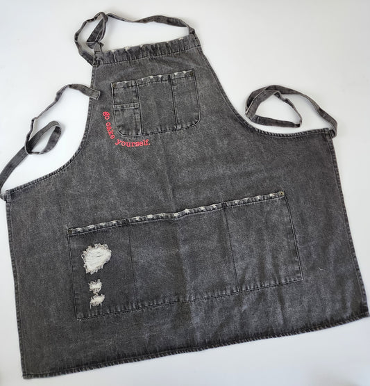 A black distressed denim apron has a utility pocket at the breast and a large open pocket at the bottom. Partially outlining the breast pocket are the words "go cake yourself" in neon pink embroidery. The bottom pocket has extra distressing in one corner for an almost torn look. Long thin straps are attached to either side of the apron, as well as a neck strap at the top secured by a brass-colored loop buckle. This apron has a classic look with punk details.