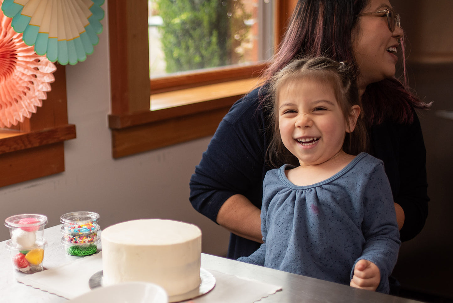 Happy little girl at a custom DIY cake decorating party in the Portland area, smiling big with an undecorated cake, sprinkles, and fondant in front of her. Ready to begin her DIY cake decorating, as her mom sits smiling behind her.