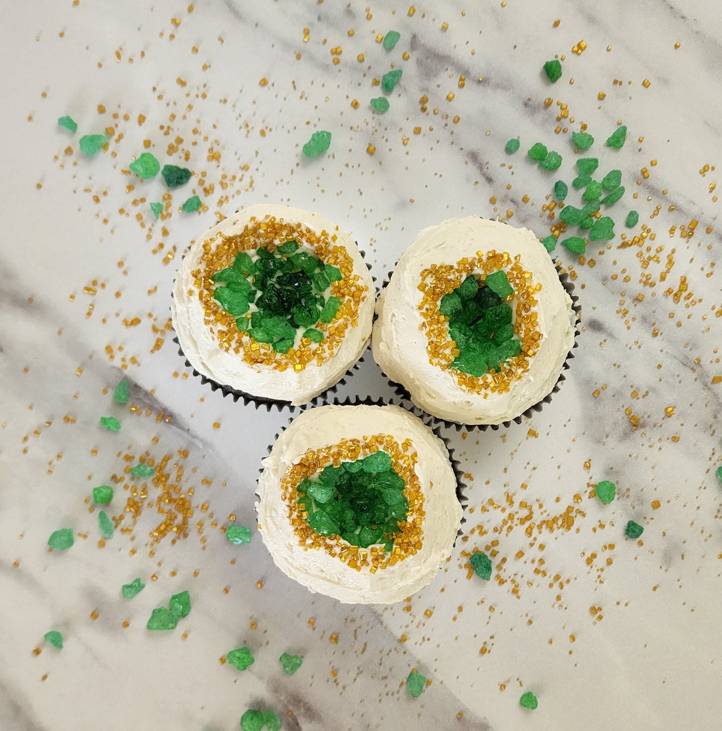 Three finished geode cupcakes from the You’re a Gem cupcake kit. They have white frosting on top with green rock candy pressed in the center to create asymmetrical geode shapes. The green rock candy darkens to create depth in the middle. The rock candy is encircled by tiny gold sprinkles. These cupcakes are the perfect mix of science, sparkles, and sweets.