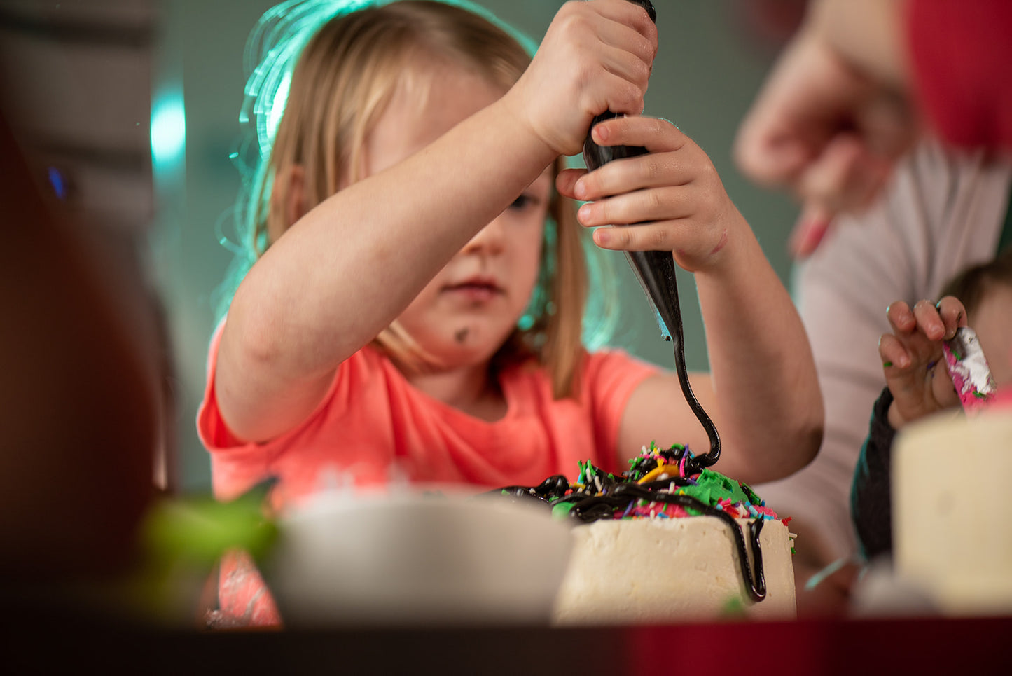 A little girl is at a DIY cake decorating party in the Portland area and is concentrating on piping frosting onto the top of her cake, where a mound of frosting and sprinkles already exists. Another out of focus kid is decorating their cake with a spatula.