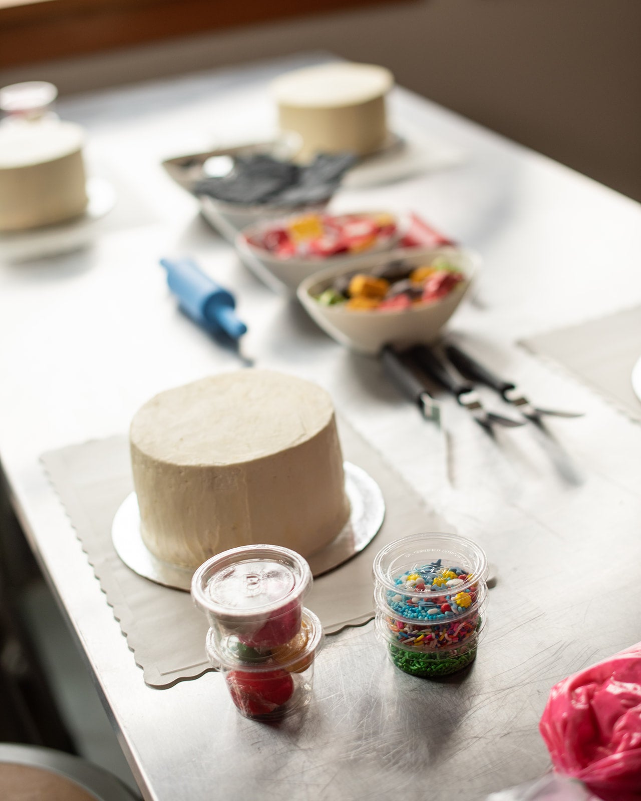  A colorful scene from a custom DIY cake decorating party in the Portland area before the decorating begins, including several frosted white cakes lying on parchment placemats, cake-decorating tools, sprinkle, and fondant.