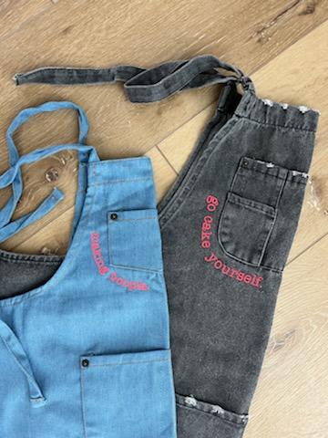 A light blue denim kid's apron folded in half rests on top of an adult's distressed black denim apron, which is also folded in half. Both adults and kids can wear custom-embroidered Cake Hoopla aprons.
