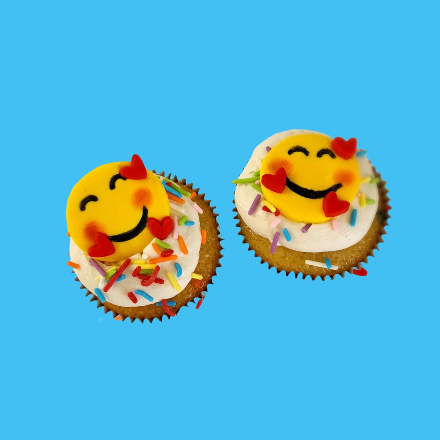 Emoji cupcakes frosted white with rainbow sprinkles and yellow smiling emoji faces with pink cheeks and three floating hearts; pre-decorated cupcakes available for shipping to the continental US.