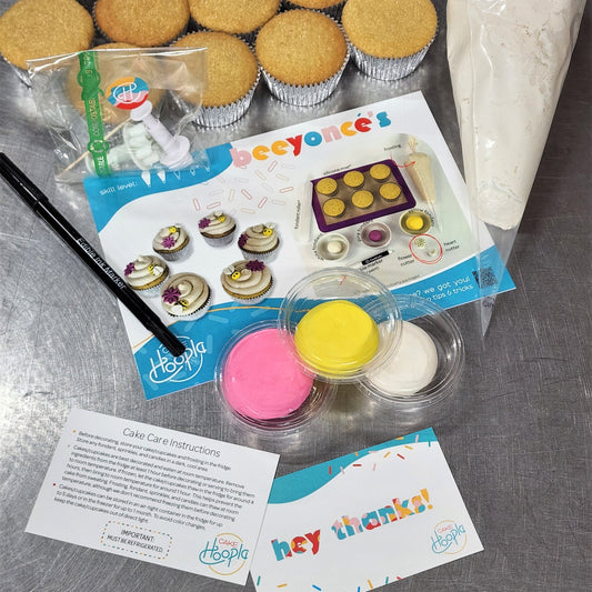 The ingredients and materials in a DIY Beeyonce's cupcake kit. 12 cupcakes, a large piping bag of white frosting, an edible black marker, pink, yellow, and white fondant, two fondant cutters in a compostable cellophane bag, a colorful instruction card with pictures, a cake care instruction card, and a thank you card.