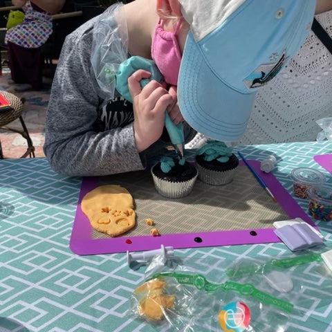 A little girl is at a custom DIY cupcake decorating party in the Portland area. She is concentrating as she pipes blue frosting onto one of two cupcakes. Around her are more decorating tools and ingredients.