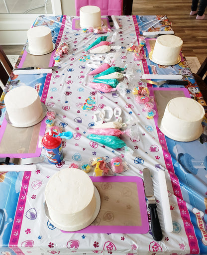 The setup for a custom dog-themed kids' cake decorating party the Portland area. A long table with bright Paw Patrol decorations contains white frosted cakes, piping bags with frosting, sprinkles, fondant, and tools for DIY cake decorating.