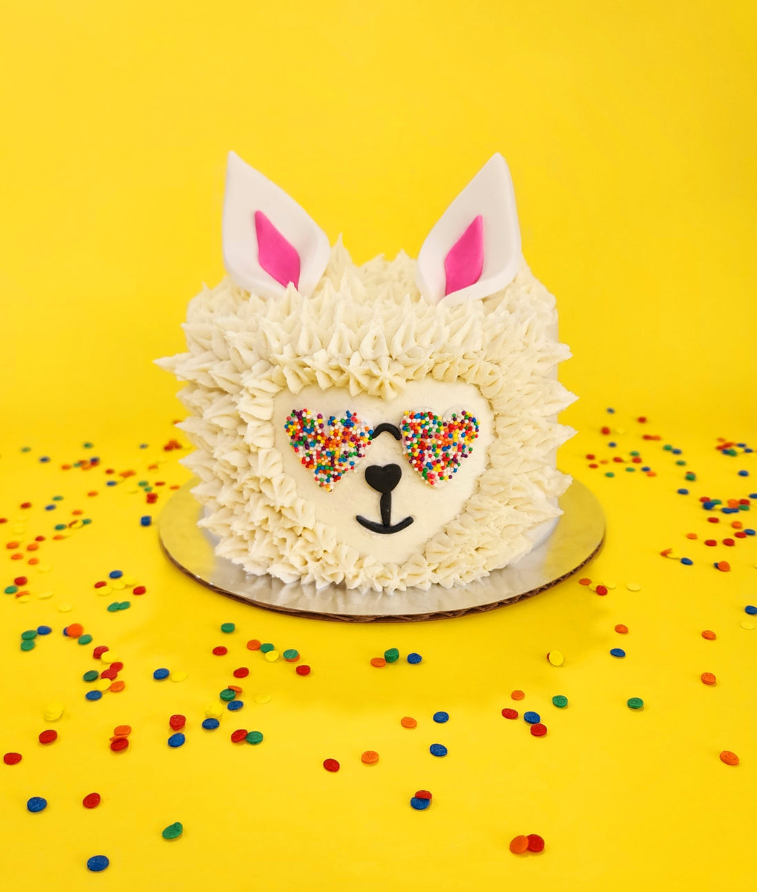 Cute llama cake piped with white frosting hair, pink and black fondant details, and heart-shaped sunglasses covered in rainbow sprinkles.
