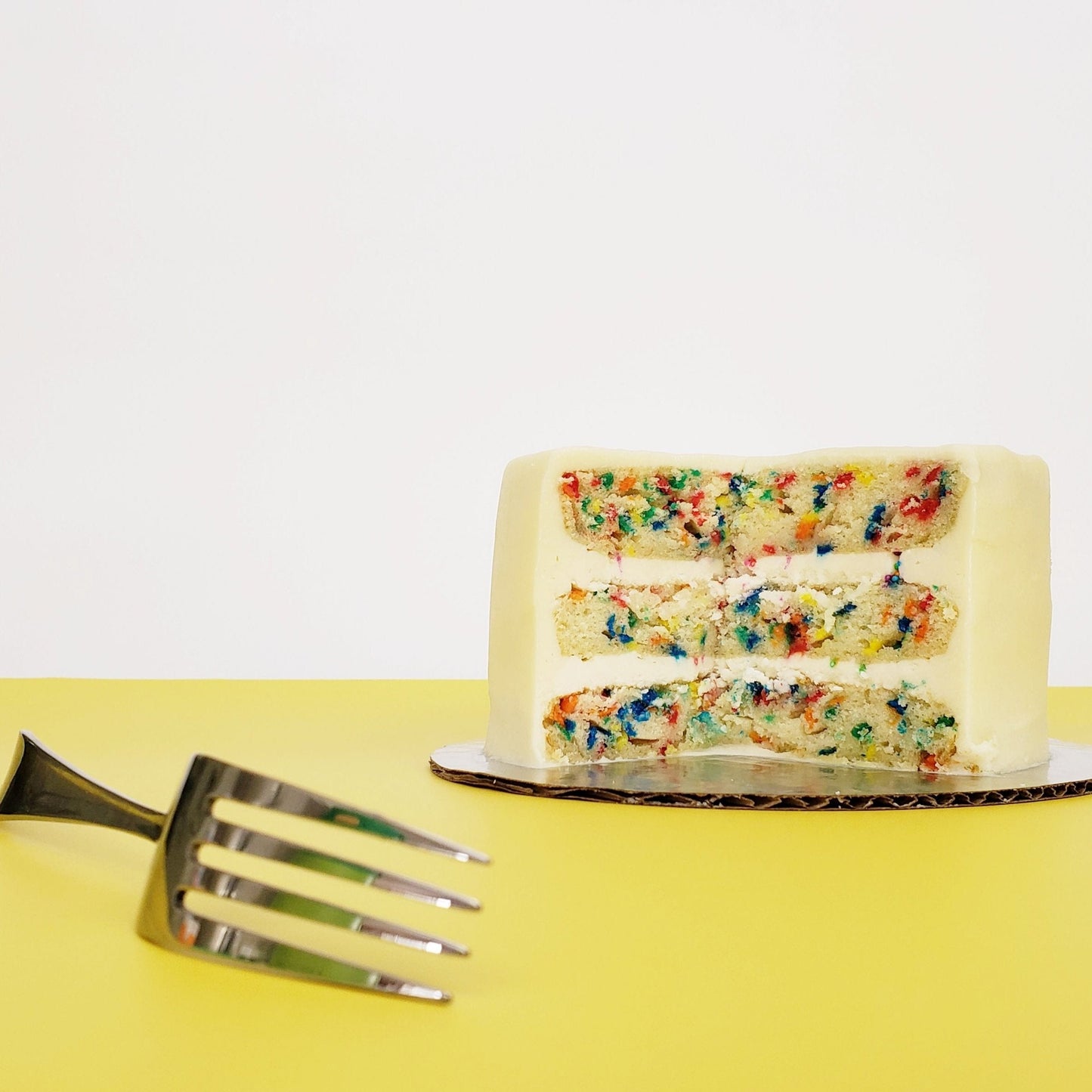 Cake Hoopla’s “Fetti” cake, cut open to reveal the inside layers. A three-layer vanilla cake has tons of bright-colored confetti sprinkles baked into the layers, and white vanilla buttercream frosting separating each layer. The cake looks moist and delicious. 