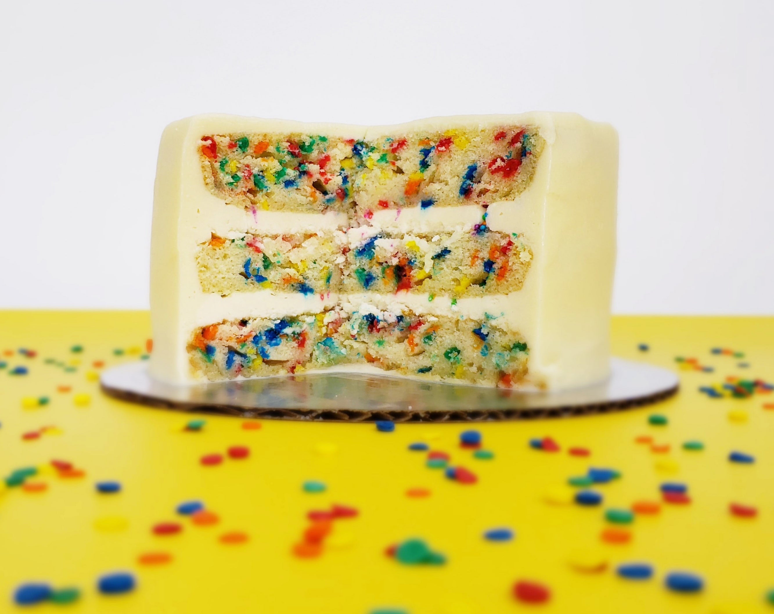 A “Fetti” cake, cut open to reveal the inside layers. A three-layer vanilla cake has tons of bright-colored confetti sprinkles baked into the layers, and white vanilla buttercream frosting separating each layer. The cake looks moist and delicious.