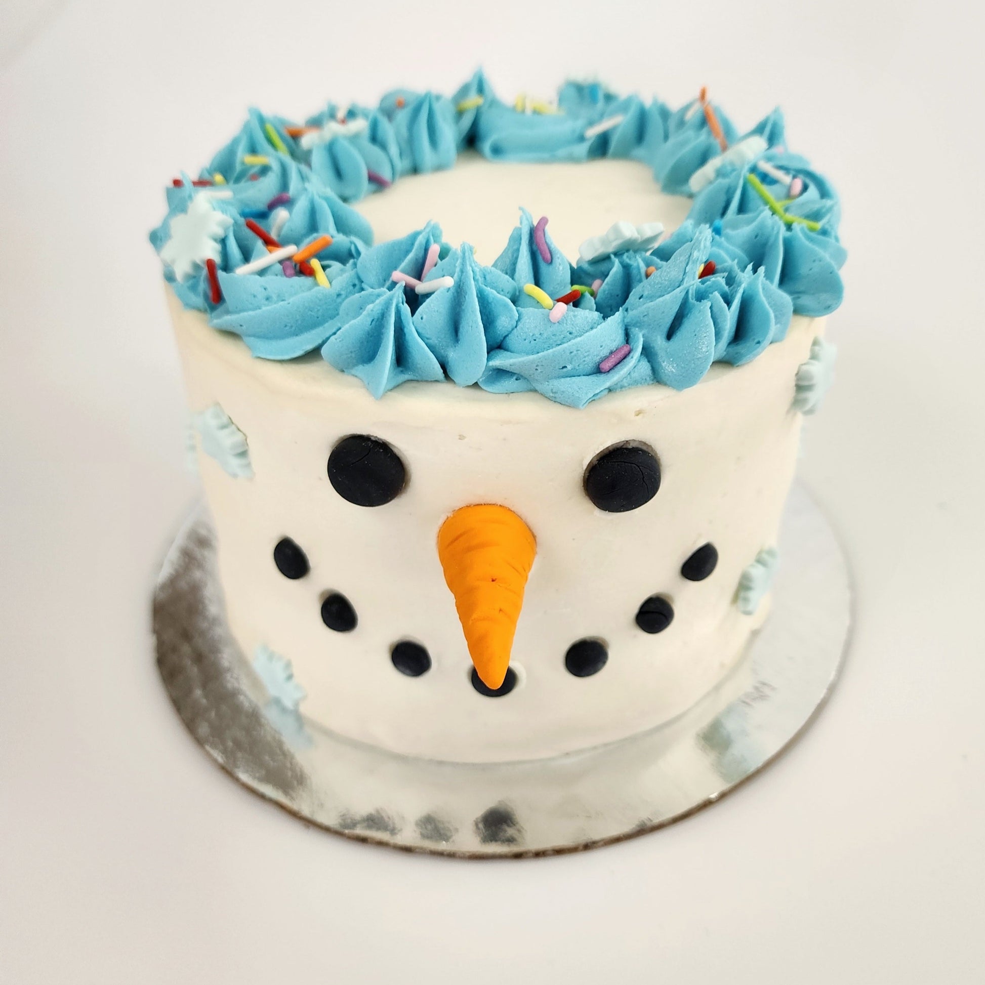 DIY cake decorating. Snowman, snowperson cake with face made out of black fondant and orange fondant nose.  Blue frosting crown topped with rainbow sprinkles and light blue snowflakes made of fondant.