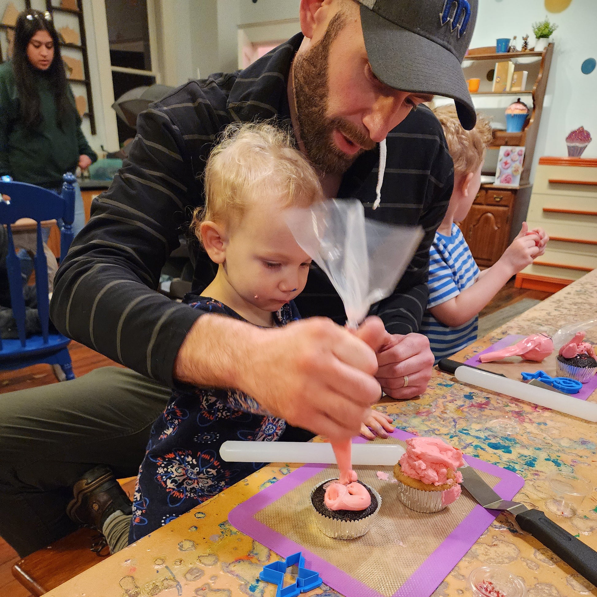 A father and toddler daughter decorate a cupcake together with pink frosting and heart sprinkles at a Cake Hoopla Valentine's Day event.