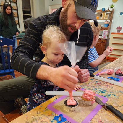 A father and toddler daughter decorate a cupcake together with pink frosting and heart sprinkles at a Cake Hoopla Valentine's Day event.