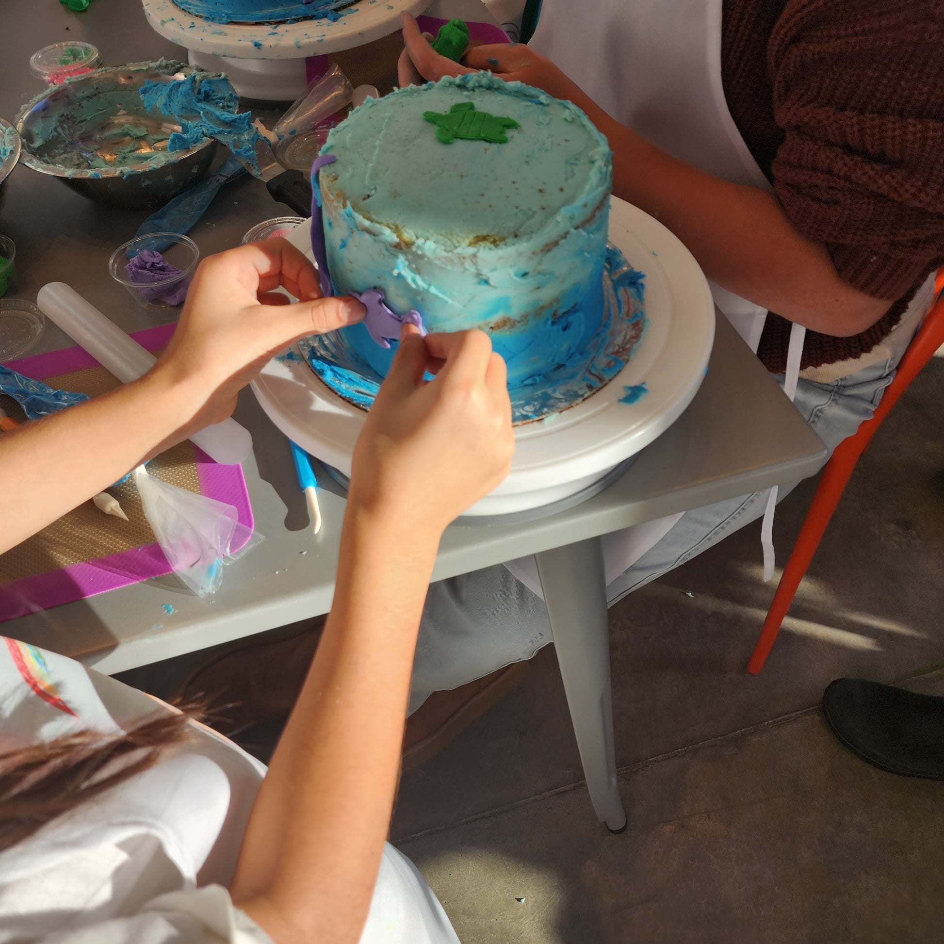 In the Cake Hoopla DIY cake/cupcake-decorating studio, after learning to frost the cake, two kids work on adding ocean-themed decorations to their ombre blue cakes, including purple fish and green turtles.