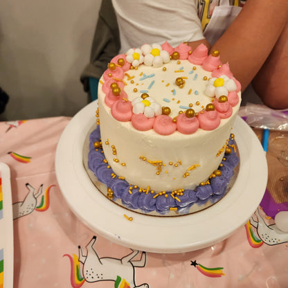 A kid displays their lovely pink, gold, purple, and white cake they learned to decorate at Cake Hoopla, a DIY cake/cupcake-decorating studio.