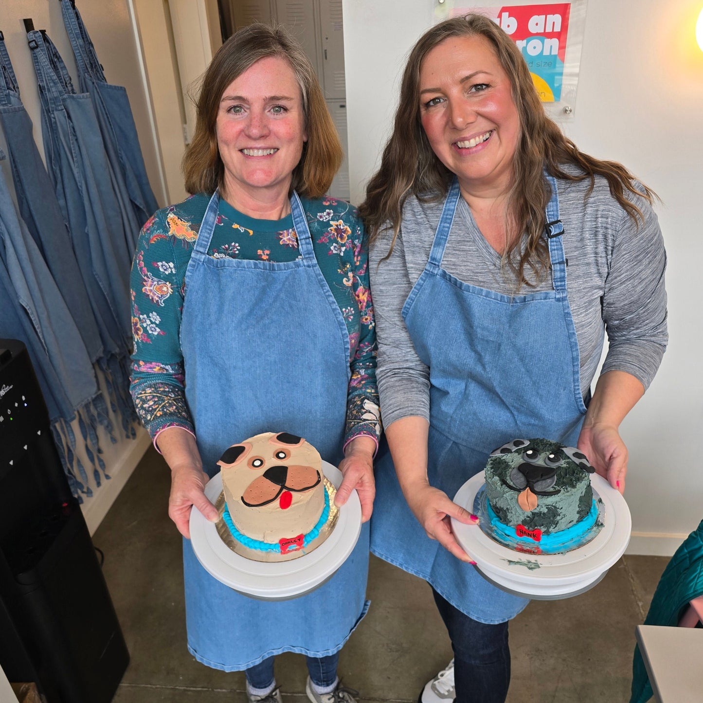 Two women proudly display their dog cakes, one brown and one silver, both with blue collars and red name tags. These were decorated a DIY cake-decorating workshop at Cake Hoopla.