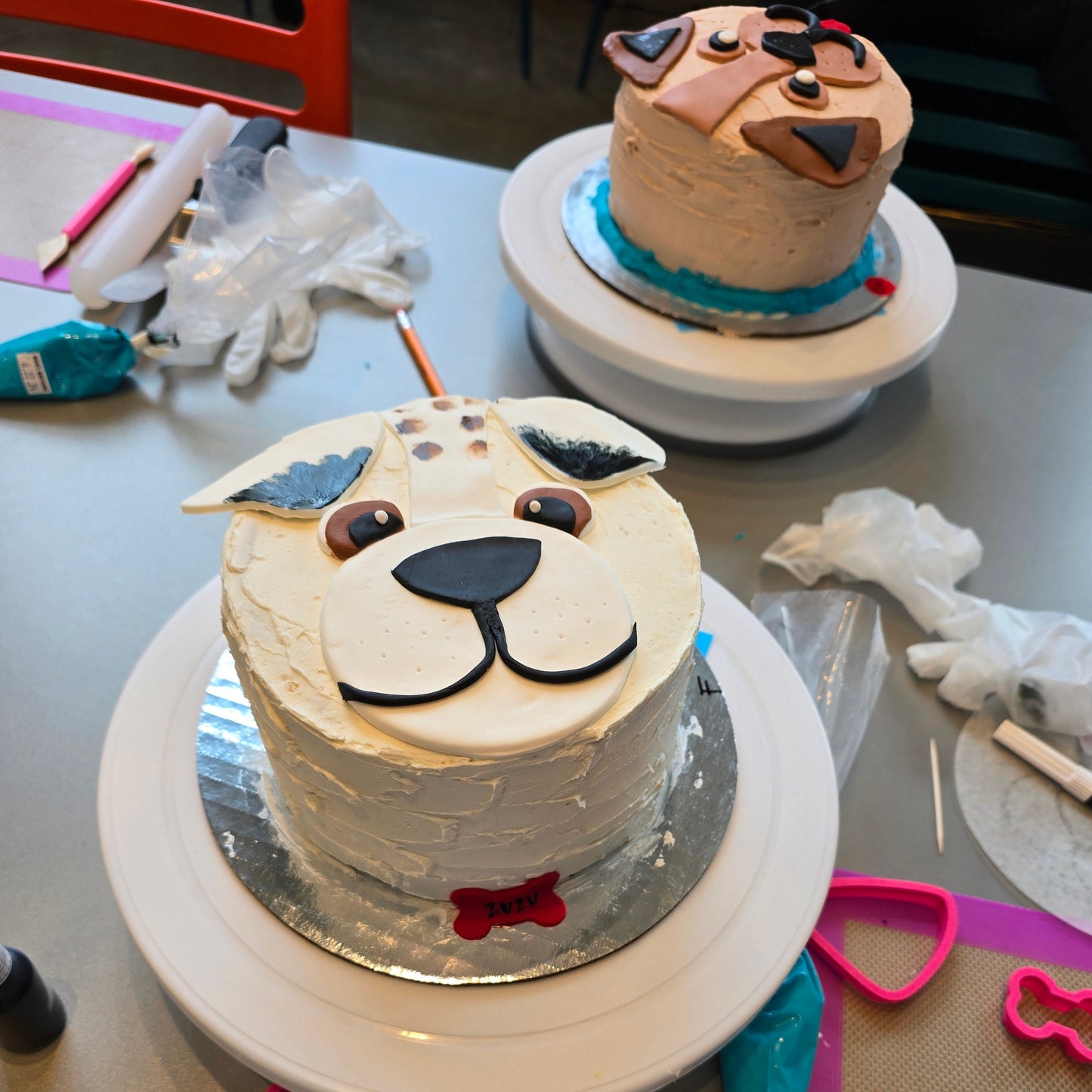 A white dog cake with the tips of its ears painted black, brown eyes, and a red bone name tag. Decorated by an attendee at a do-it-yourself cake-decorating workshop at Cake Hoopla.
