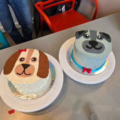 Two cute dog cakes, one gray with blue collar and one brown and white with a red bow, both decorated during a do-it-yourself cake-decorating workshop.