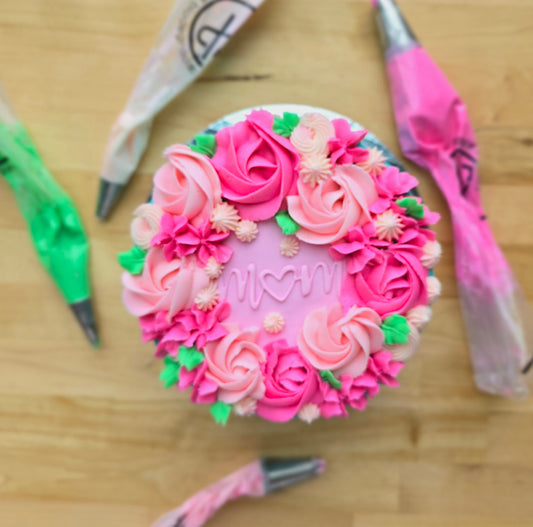 A bright floral-themed cake available during a Mother's Day workshop, with pink rosettes, green leaves, and mom written in the center. 