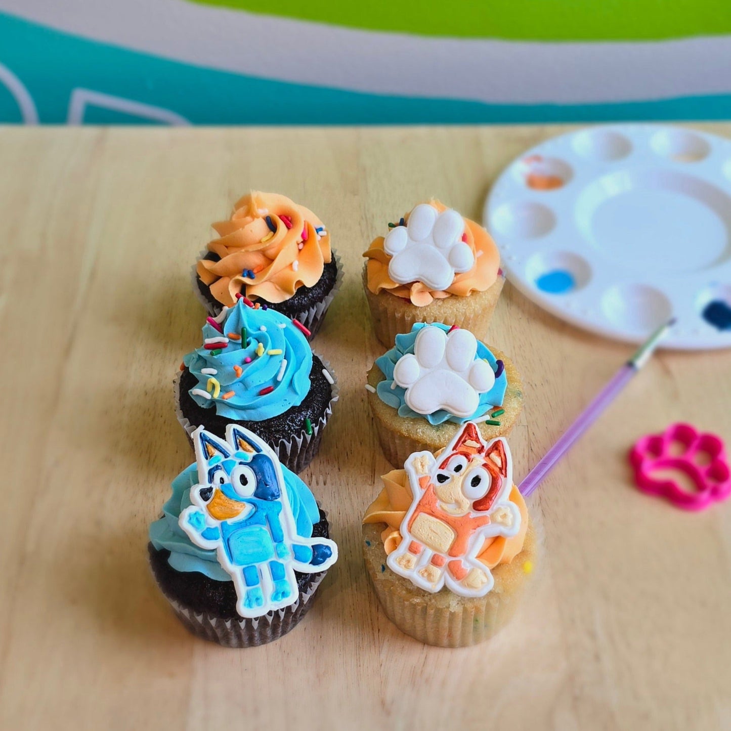 Cupcakes decorated during a DIY cupcake-decorating workshop at Cake Hoopla with blue and orange frosting, fondant dog paws, and blue and red heeler dogs who look like the cartoon characters Bluey and Bingo