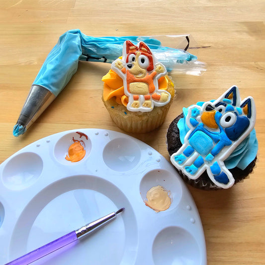 Cupcakes decorated during a DIY cupcake-decorating workshop at Cake Hoopla with blue and orange frosting, fondant dog paws, and blue and red heeler dogs who look like the cartoon characters Bluey and Bingo. A paint pallet and paintbrush and piping bag of blue frosting are nearby.