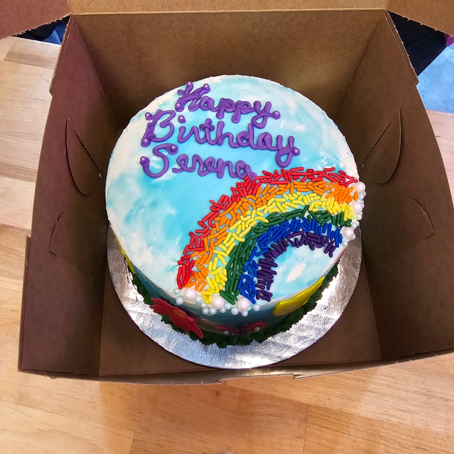 A birthday cake frosted with white and blue frosting, pink fondant flowers, and rainbow sprinkles from a Cake Hoopla Create Your Own Cake Kit available during  time, as a to-go kit, or as a shipped kit from Cake Hoopla in Tigard, Oregon near Portland.