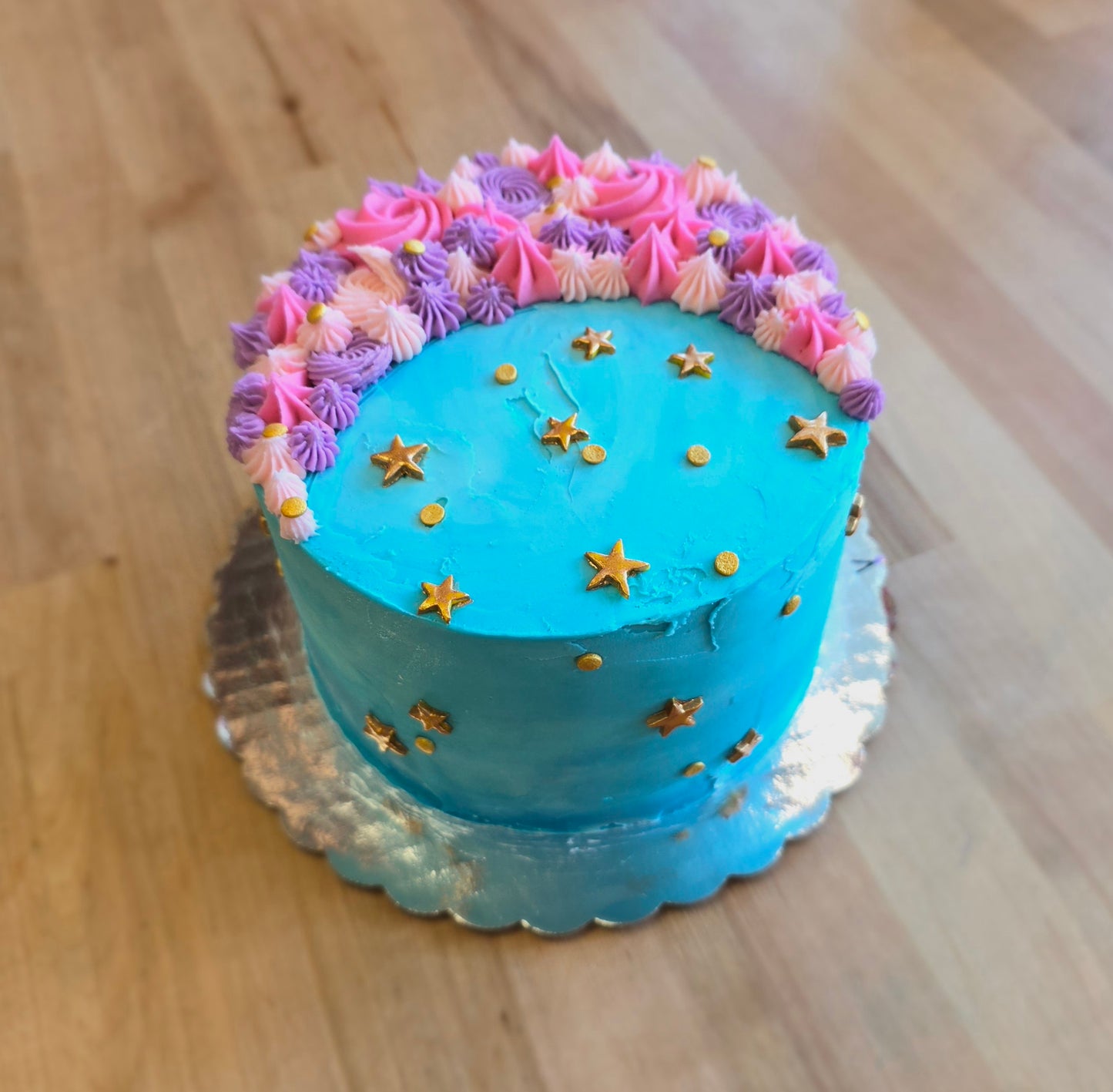 A moon-themed cake frosted in teal with pink and purple crescent moon on top and gold stars dotting the cake; a pre-decorated cake available for purchase at Cake Hoopla in Tigard, Oregon.