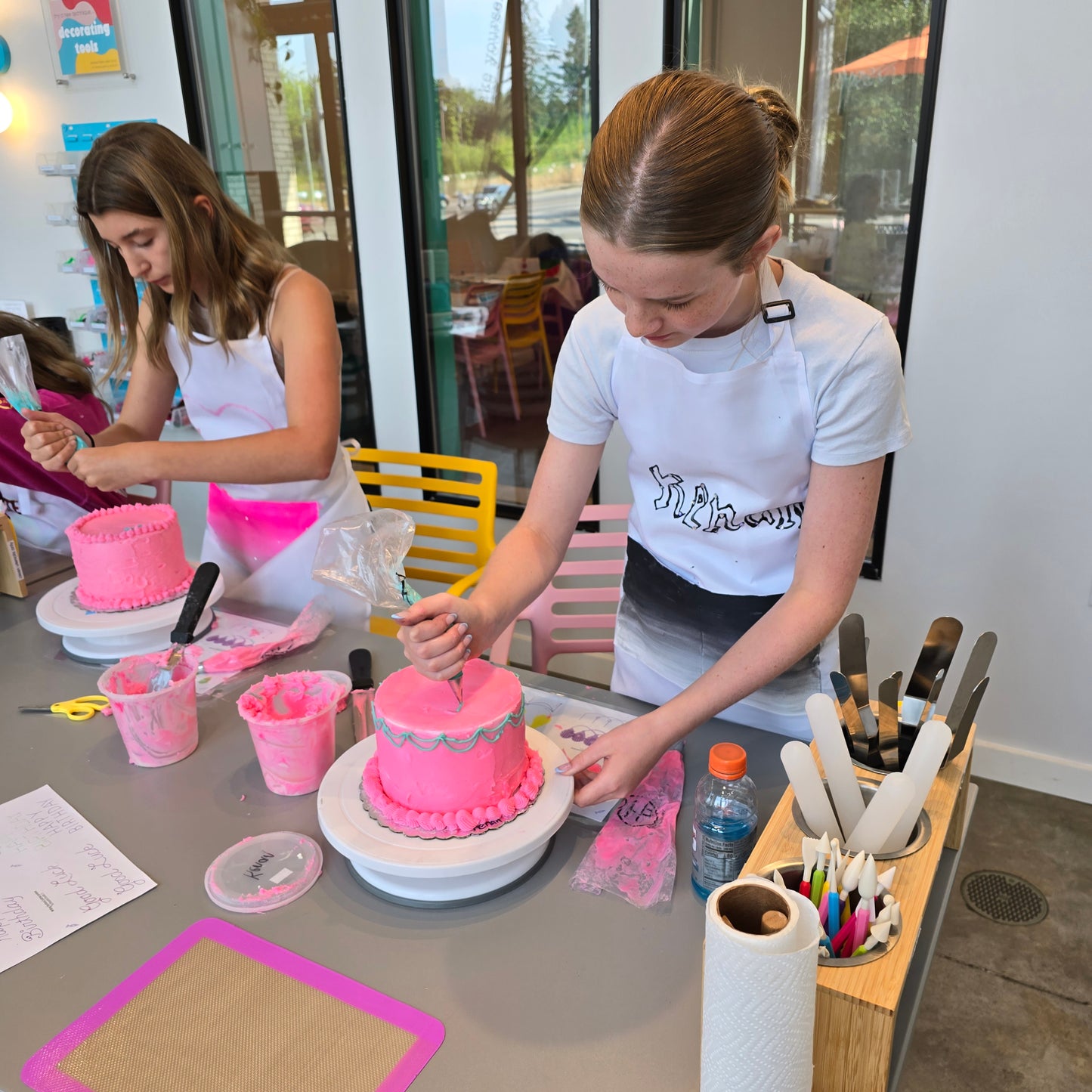 A young person works on frosting their cake pink and adding light blue drop string borders during a cake-decorating class at Cake Hoopla in Tigard, Oregon near Portland.