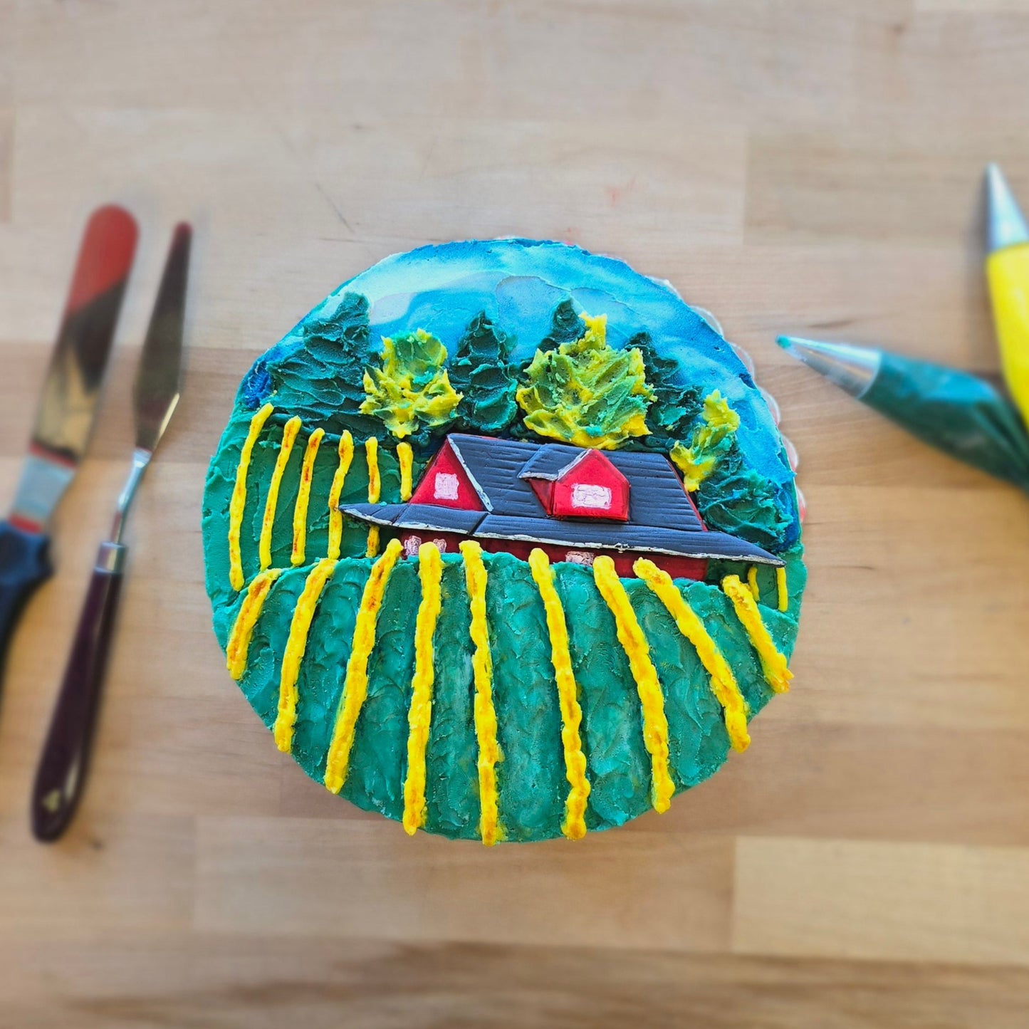 Round cake decorated during fall-themed DIY cake-decorating workshop, where top has rows of yellow grape vines, Sokol Blosser's big read house, and trees against a blue sky background.