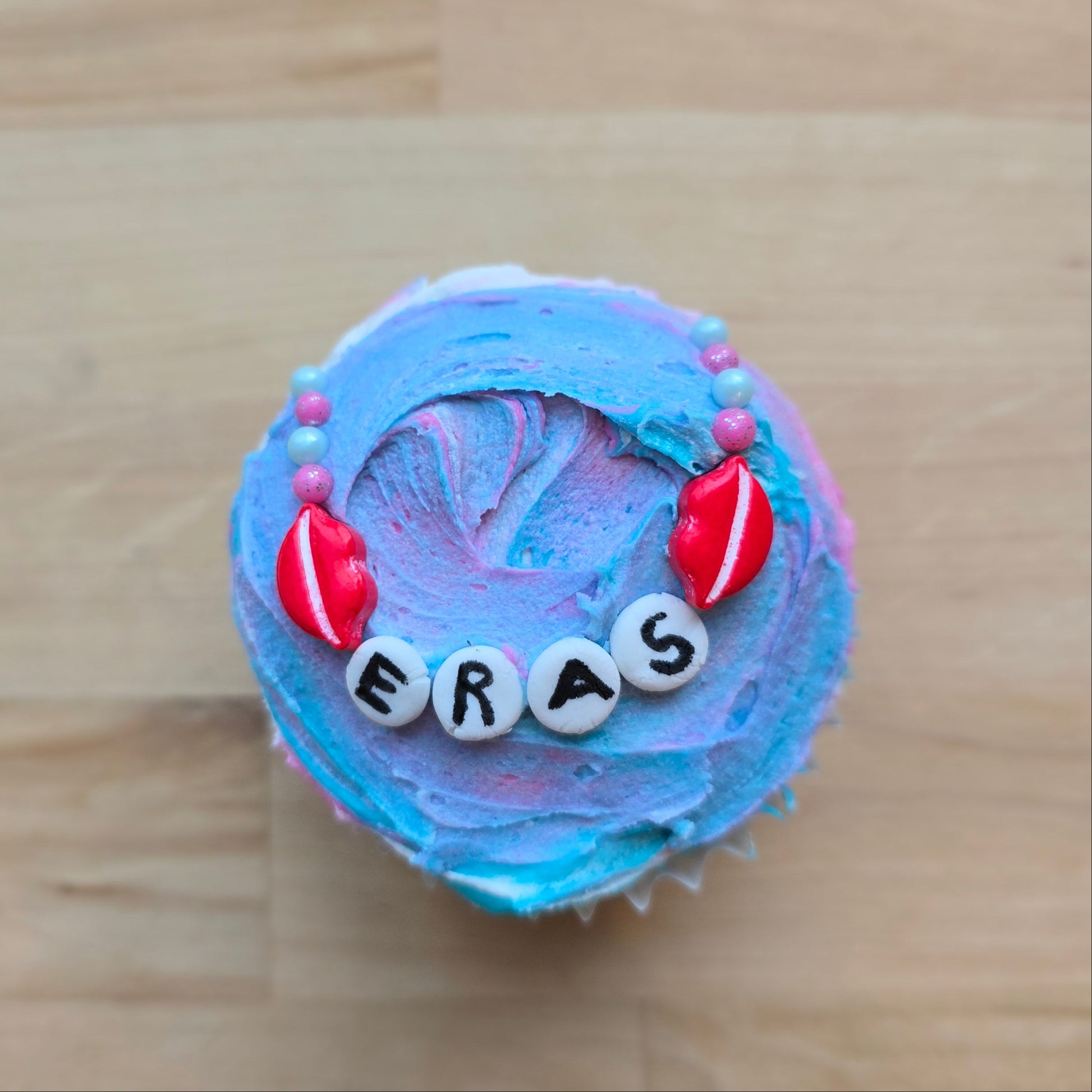 Friendship-bracelet-themed cupcake decorated at Cake Hoopla in Tigard, Oregon near Portland as a DIY cupcake-decorating kit, with the word "ERAS" written on it.