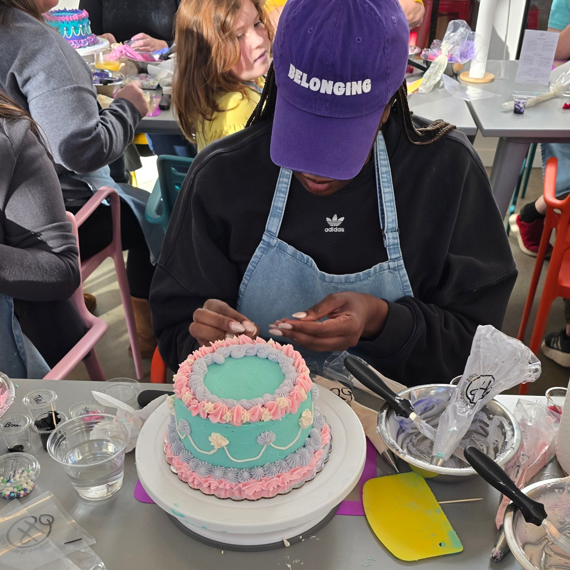 A woman decorates a a purple, teal, and pink cake with piped borders, shells, and writing at Cake Hoopla in Tigard, Oregon, near Portland, during a cake-decorating class. 