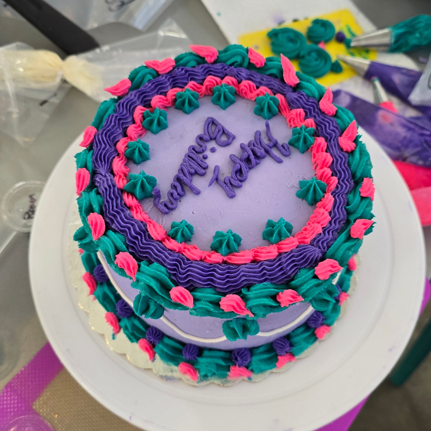 A purple, teal, and pink cake decorated with piped borders, shells, and writing at Cake Hoopla in Tigard, Oregon, near Portland, during a cake-decorating class. 