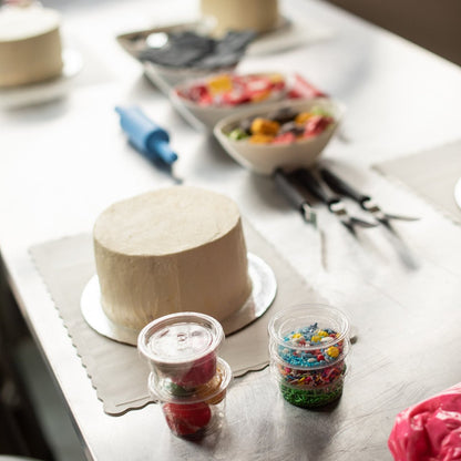  A colorful scene from a custom DIY cake decorating party in the Portland area before the decorating begins, including several frosted white cakes lying on parchment placemats, cake-decorating tools, sprinkle, and fondant.