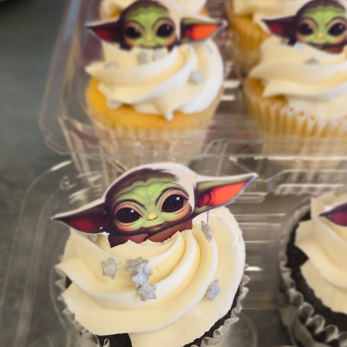 Frosted cupcakes topped with a small Star Wars Grogu/Baby Yoda topper, arranged in a clear plastic cupcake tray.