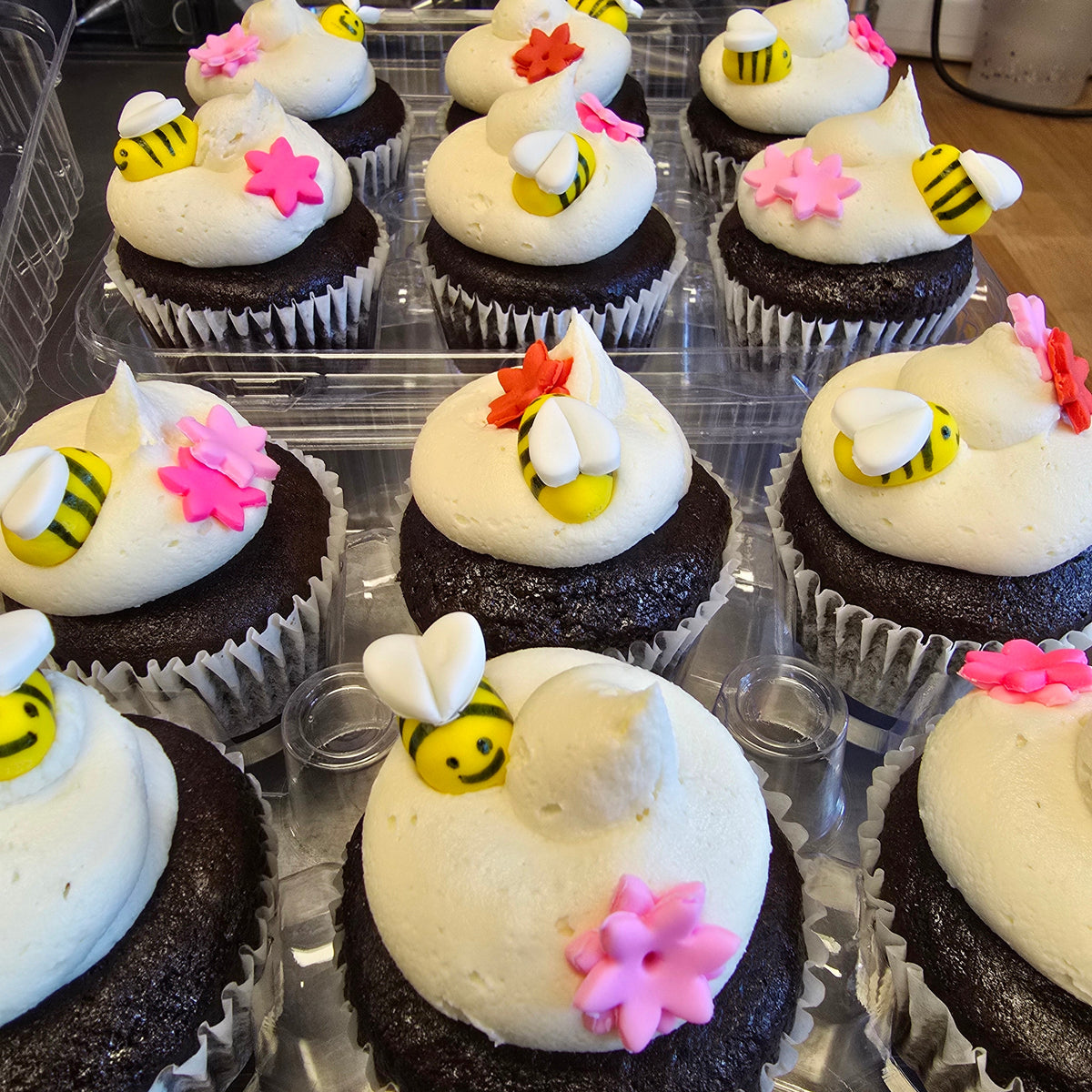 A tray of chocolate cupcakes decorated with white frosting, bumblebee toppers, pink flower accents, and small yellow-and-white bee details, photographed in a retail or bakery setting.