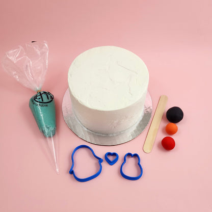 The components of the "I Flippin' Love You" DIY cake-decorating kit, including a white frosted cake, piping bag with teal frosting, three colors of fondant, a wood spatula, and penguin and heart fondant cutters.