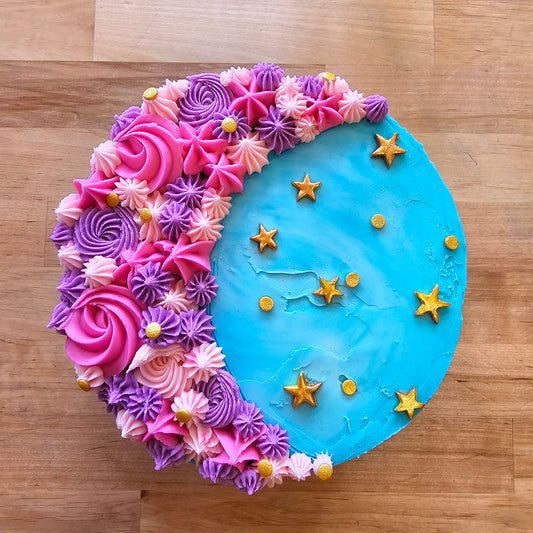 A decorated cake shaped like a crescent moon, with one half covered in pink, purple, and light pink piped frosting rosettes and stars, and the other half in bright blue frosting decorated with small gold star sprinkles, shown on a wooden surface.