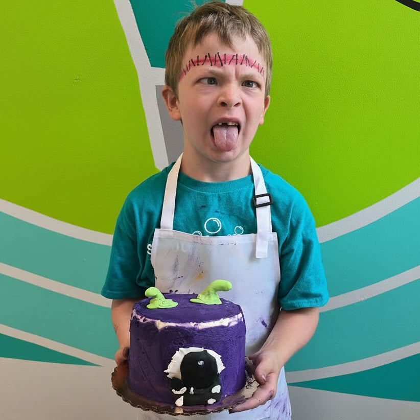 Boy holds his purple monster cake in front of a colorful background, displaying a silly face and face painted stiches across his forehead.