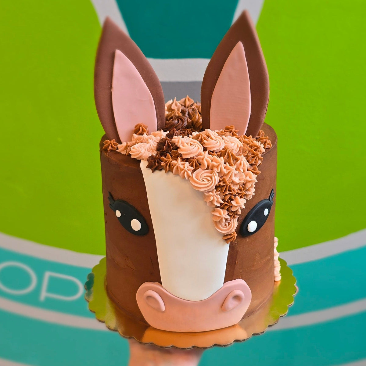 A person's hand holding a small horse-shaped cake decorated with brown frosting, pink ears, and a white blaze on the forehead, set against a bright green and teal geometric wall background.