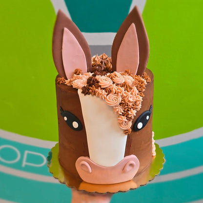 A person's hand holding a small horse-shaped cake decorated with brown frosting, pink ears, and a white blaze on the forehead, set against a bright green and teal geometric wall background.